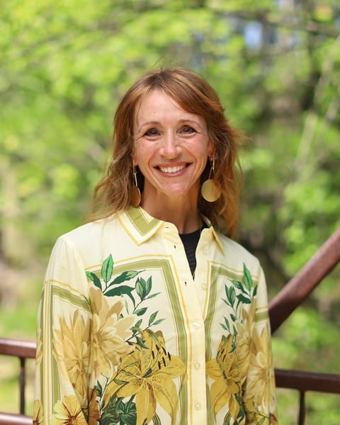 Woman with reddish-brown hair smiling outdoors, wearing a yellow floral button-up shirt with green foliage designs